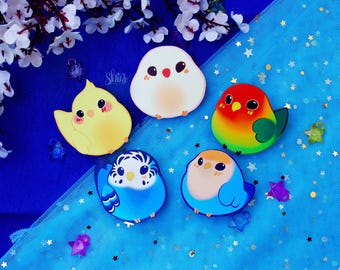 May include: Five colorful enamel pins featuring different types of birds. The pins are on a blue background with white stars.