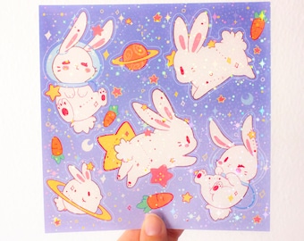 May include: A sheet of holographic stickers featuring five white bunnies in various poses, including one wearing a space helmet. The stickers are set against a purple background with glitter and stars. There are also orange planets and carrots on the sheet.