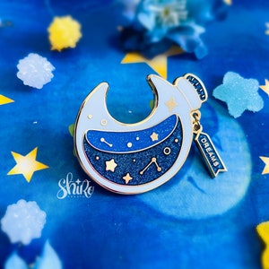 Puede incluir: Un pin de esmalte dorado en forma de media luna con un cielo estrellado azul y blanco en su interior. El pin tiene una pequeña etiqueta que dice "DREAMS" colgando de él.