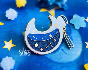 May include: A gold-tone enamel pin shaped like a crescent moon with a blue and white starry sky inside. The pin has a small tag that says "DREAMS" hanging from it.