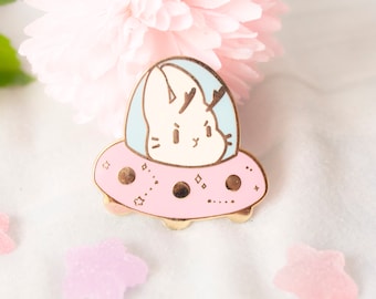 May include: A gold-colored enamel pin featuring a white bunny with antlers inside a pink and blue spaceship. The spaceship has gold accents and a dotted pattern.
