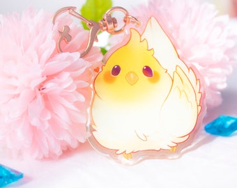 May include: A yellow and white cartoon chick keychain with a pink heart on its cheek. The keychain is made of clear acrylic and has a gold metal ring.