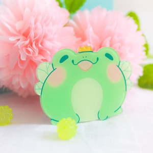 May include: A green frog with a crown and wings, made of acrylic. The frog is smiling and has a pink blush on its cheeks. The frog is sitting on a white surface with two yellow, bumpy balls.