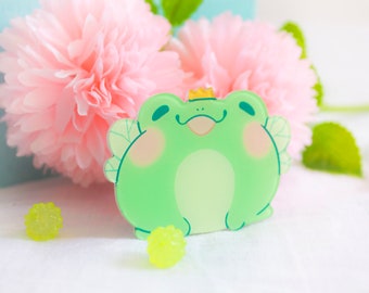 May include: A green frog with a crown and wings, made of acrylic. The frog is smiling and has a pink blush on its cheeks. The frog is sitting on a white surface with two yellow, bumpy balls.