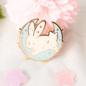 Könnte beinhalten: Ein goldfarbener Metallpin mit einem hellblauen Halbmond und einem weißen Hasen, der darüber springt. Der Hase hat lange Ohren und ist mit kleinen Diamanten übersät. Der Pin ist ein niedliches, skurriles Accessoire.