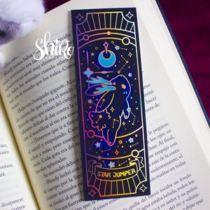 Può includere: Un segnalibro nero con un design olografico raffigurante un coniglio, stelle e una luna crescente. Il testo "STAR JUMPER" è visualizzato su uno striscione. Il segnalibro è posto in un libro aperto.