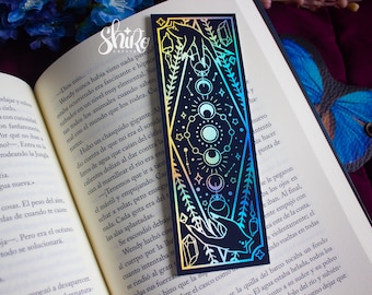 May include: A black bookmark with a holographic finish features a hand holding a crystal with a crescent moon and stars. The design is surrounded by a diamond shape with floral accents.