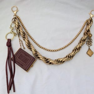 Könnte beinhalten: Ein goldfarbenes Accessoire mit mehreren Kettensträngen, bestehend aus einer dicken gedrehten Gliederkette, einer feinen Perlenkette und einer kleineren Kette. Es enthält eine dunkelburgunderfarbene Lederquaste und ein kleines Lederetikett.