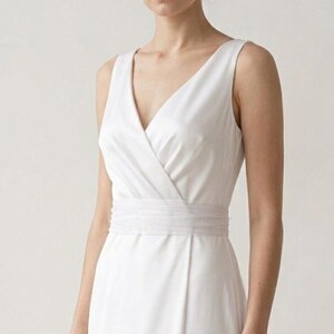 Könnte beinhalten: Ein ärmelloses weißes Kleid mit V-Ausschnitt und Wickel-Oberteil. Das Kleid hat eine taillierte Taille mit einem strukturierten Band und einen fließenden Rock. Das Kleid ist aus einem glatten, blickdichten Stoff gefertigt.