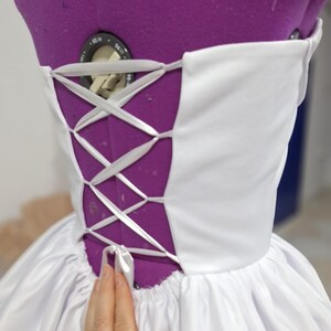 May include: A white corset-style bodice with a lace-up back detail. The back is a deep purple color, and the white fabric is gathered at the waist. The garment is on a purple dress form.