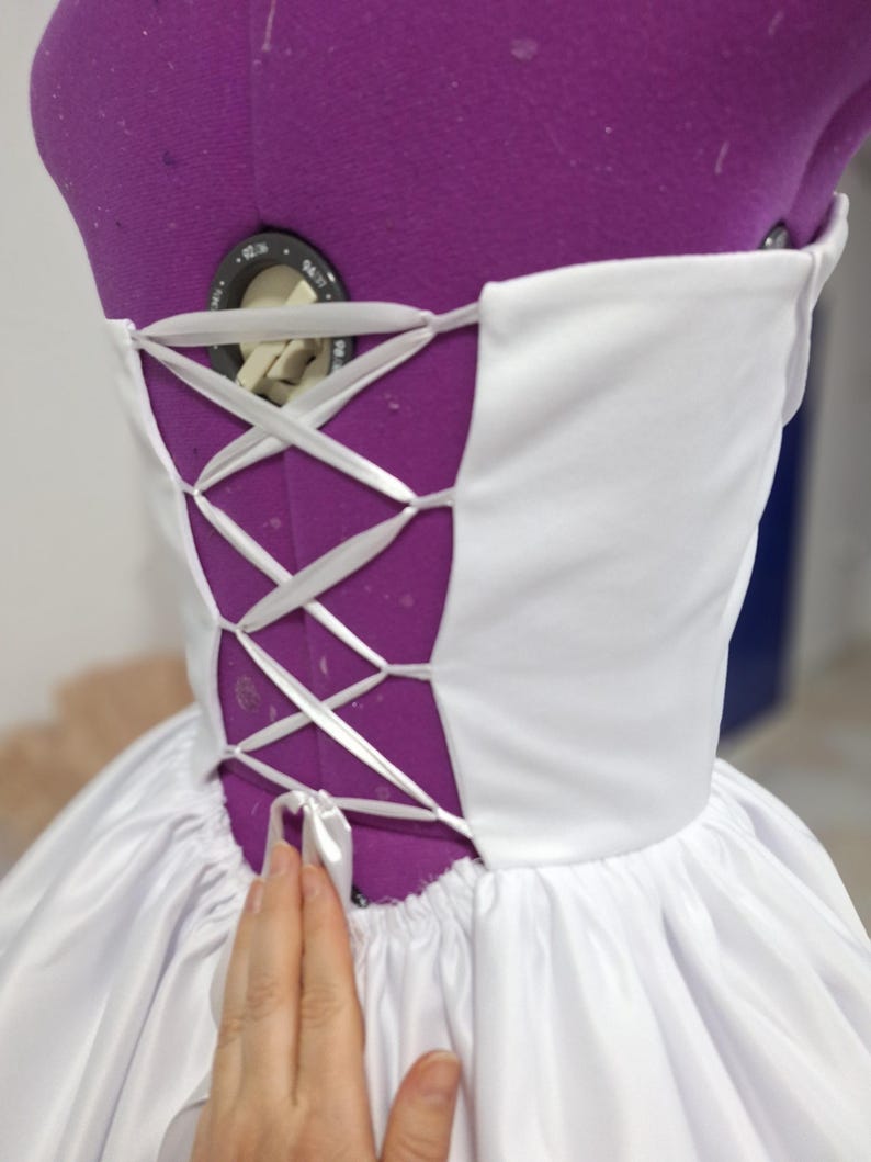 May include: A white corset-style bodice with a lace-up back detail. The back is a deep purple color, and the white fabric is gathered at the waist. The garment is on a purple dress form.