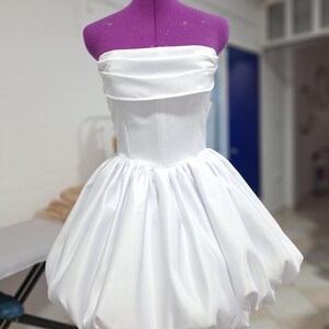 May include: A white strapless dress with a fitted bodice and a voluminous, bubble-shaped skirt. The dress is displayed on a purple dress form. The fabric appears to be a smooth, satin-like material.
