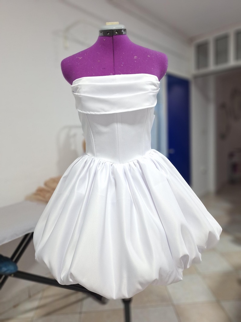 May include: A white strapless dress with a fitted bodice and a voluminous, bubble-shaped skirt. The dress is displayed on a purple dress form. The fabric appears to be a smooth, satin-like material.