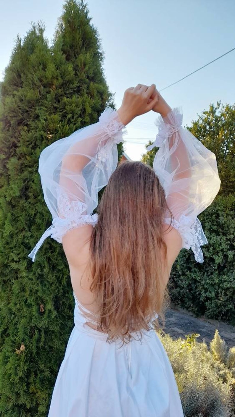 K&ouml;nnte beinhalten: Ein wei&szlig;es, schulterfreies Kleid mit durchsichtigen, mit Spitze besetzten &Auml;rmeln. Das Kleid hat ein tailliertes Oberteil und einen flie&szlig;enden Rock.