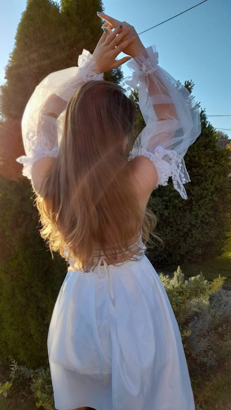 K&ouml;nnte beinhalten: Ein wei&szlig;es Kleid mit durchsichtigen, mit Spitze besetzten &Auml;rmeln. Das Kleid hat ein tailliertes Oberteil und einen weiten Rock. Das Model tr&auml;gt das Kleid mit offenem Haar und steht in einem Garten.