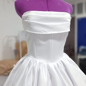 May include: A white strapless dress on a purple dress form. The bodice is fitted with vertical seams, and the skirt is full and gathered. The dress appears to be made of a satin-like fabric, and the overall style is elegant and formal.