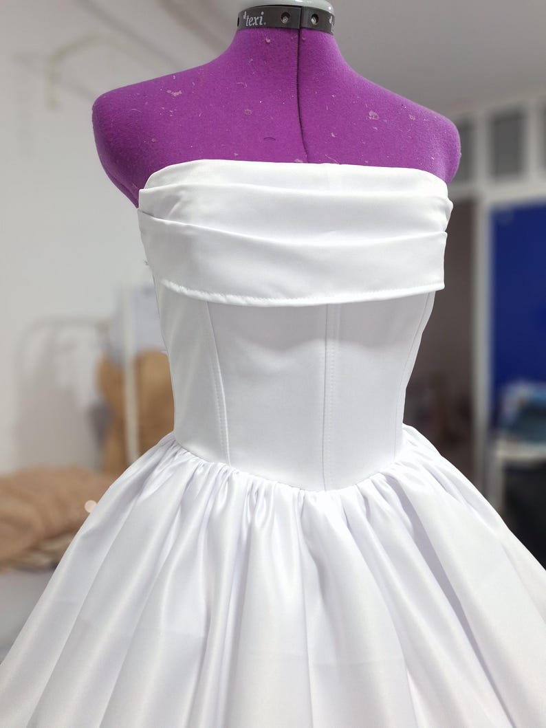 May include: A white strapless dress on a purple dress form. The bodice is fitted with vertical seams, and the skirt is full and gathered. The dress appears to be made of a satin-like fabric, and the overall style is elegant and formal.
