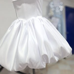 May include: A white satin dress with a fitted bodice and a bubble skirt. The dress is displayed on a mannequin. The bodice has a straight neckline and a structured design. The skirt is full and gathered, creating a voluminous shape. The dress is a classic design.
