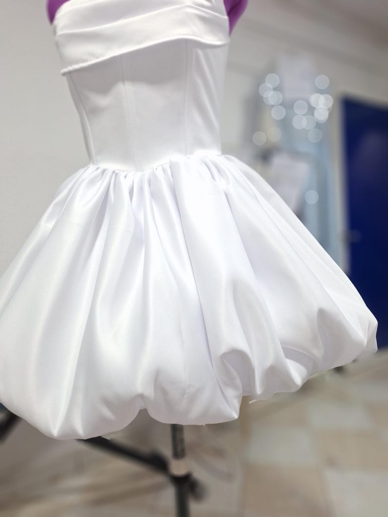 May include: A white satin dress with a fitted bodice and a bubble skirt. The dress is displayed on a mannequin. The bodice has a straight neckline and a structured design. The skirt is full and gathered, creating a voluminous shape. The dress is a classic design.