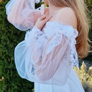 K&ouml;nnte beinhalten: Wei&szlig;es, schulterfreies Kleid mit durchsichtigen Spitzen&auml;rmeln und einem R&uuml;schenkragen. Das Kleid ist aus einem leichten Stoff gefertigt und hat eine flie&szlig;ende Silhouette.