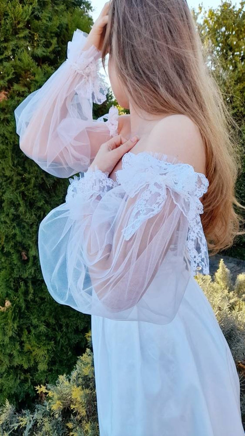 K&ouml;nnte beinhalten: Wei&szlig;es, schulterfreies Kleid mit durchsichtigen Spitzen&auml;rmeln und einem R&uuml;schenkragen. Das Kleid ist aus einem leichten Stoff gefertigt und hat eine flie&szlig;ende Silhouette.