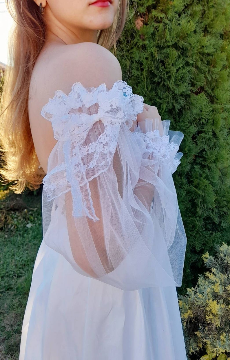 K&ouml;nnte beinhalten: Wei&szlig;es, schulterfreies Kleid mit durchsichtigen Spitzen&auml;rmeln und einem Schleifendetail.