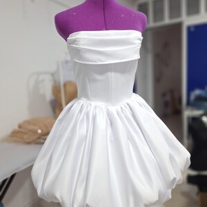 May include: A white strapless dress with a fitted bodice and a voluminous, bubble-shaped skirt. The dress is displayed on a purple dress form. The fabric has a slight sheen, suggesting a satin or similar material. The design is simple and elegant.