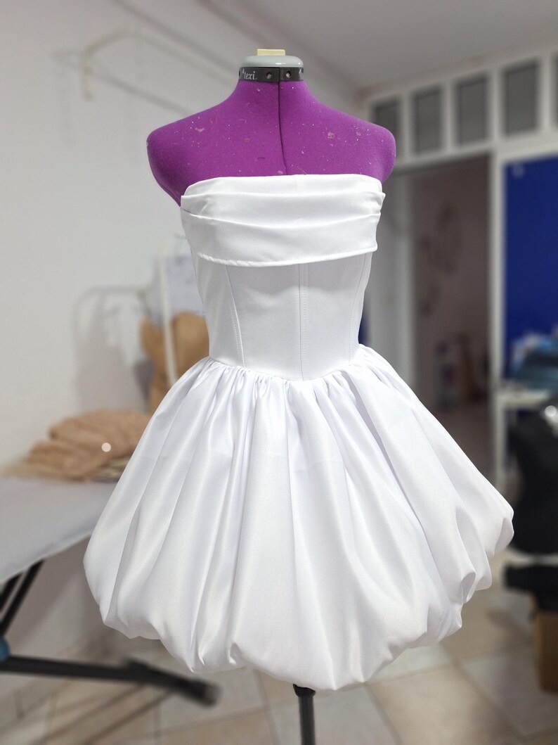 May include: A white strapless dress with a fitted bodice and a voluminous, bubble-shaped skirt. The dress is displayed on a purple dress form. The fabric has a slight sheen, suggesting a satin or similar material. The design is simple and elegant.