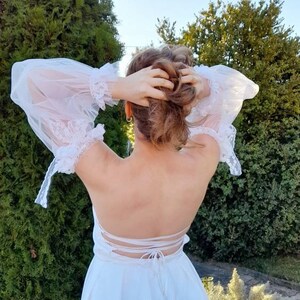 K&ouml;nnte beinhalten: Ein wei&szlig;es Kleid mit Spitzenbesatz und einem tiefen R&uuml;cken. Das Kleid ist hinten mit einem Band gebunden.