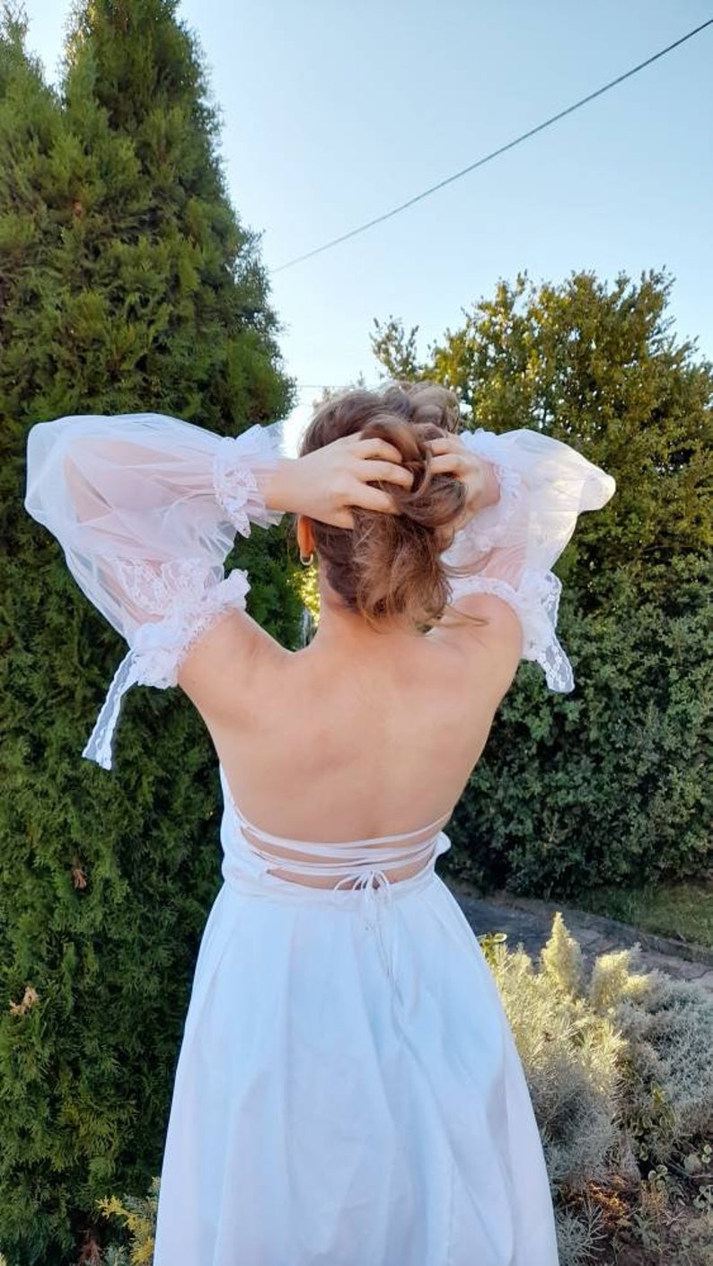 K&ouml;nnte beinhalten: Ein wei&szlig;es Kleid mit Spitzenbesatz und einem tiefen R&uuml;cken. Das Kleid ist hinten mit einem Band gebunden.
