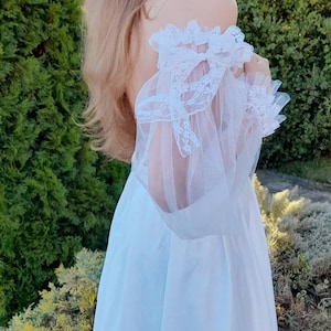 K&ouml;nnte beinhalten: Ein wei&szlig;es, schulterfreies Kleid mit einem durchsichtigen Spitzen&uuml;berzug und einem Schleifendetail an der Schulter.
