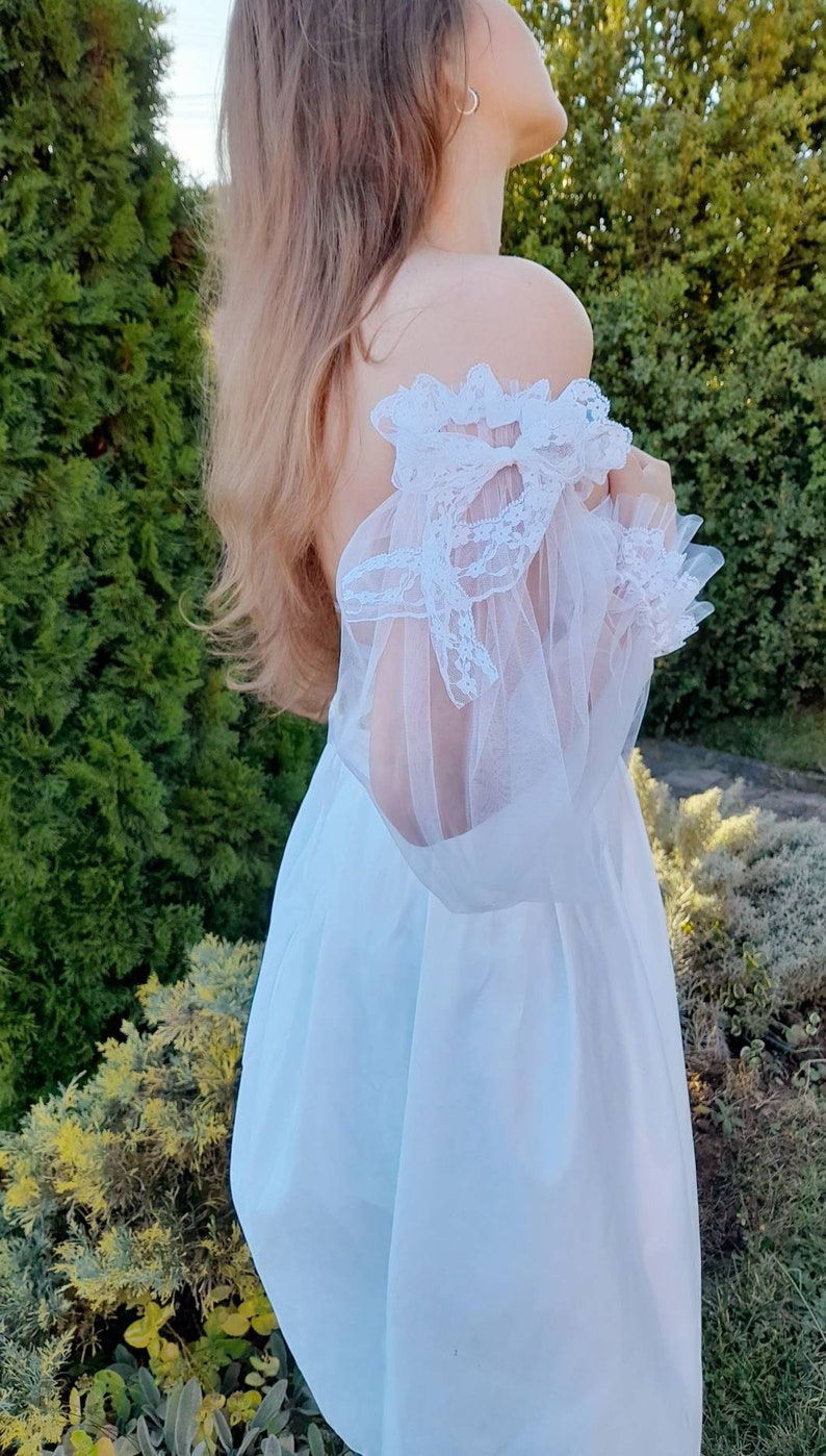 K&ouml;nnte beinhalten: Ein wei&szlig;es, schulterfreies Kleid mit einem durchsichtigen Spitzen&uuml;berzug und einem Schleifendetail an der Schulter.