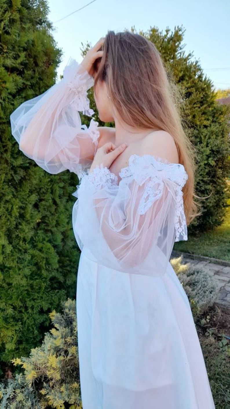 K&ouml;nnte beinhalten: Ein wei&szlig;es, schulterfreies Kleid mit durchsichtigen, mit Spitze besetzten &Auml;rmeln. Das Kleid hat einen flie&szlig;enden Rock und ein tailliertes Oberteil.