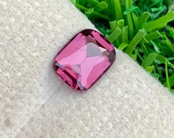 May include: A faceted, rectangular, magenta gemstone rests on a beige fabric, set against a backdrop of vibrant green artificial grass. The gemstone's cut allows light to reflect, creating a sparkling effect. The overall composition highlights the gem's color and clarity.