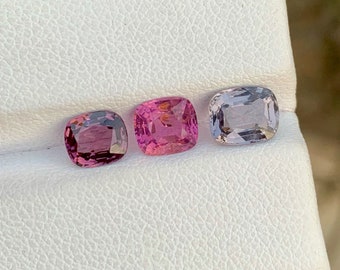 Può includere: Tre pietre preziose di forma quadrata su una superficie bianca. Le pietre preziose sono rosa, rosa chiaro e viola.