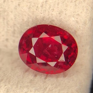 Peut inclure: Une pierre précieuse rubis de forme ovale, d'un rouge profond et présentant un motif étoilé en son centre. Elle repose sur une surface blanche et texturée, avec un fond sombre.