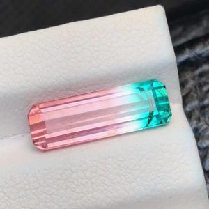 May include: A rectangular gemstone with a color gradient, transitioning from pink to teal. The gem is faceted, with visible parallel lines running along its length. The stone is set against a white surface.