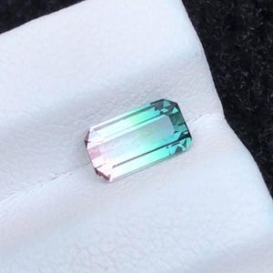 May include: A rectangular gemstone with a gradient of colors, including pink, teal, and green. The gem is set on a white surface, with a dark background. The gemstone is faceted, reflecting light and showcasing its various hues.