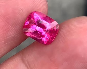 May include: A vibrant, cushion-cut, hot pink gemstone. The gem is held between fingers, showcasing its facets and brilliant color. The stone's shape is a square with rounded corners, reflecting light.