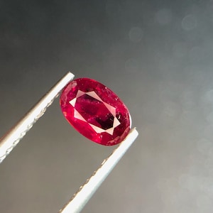 Puede incluir: Una gema ovalada de color rojo vibrante sostenida por unas pinzas sobre un fondo gris borroso.