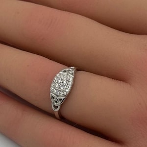 May include: A silver ring with a square, diamond-studded center stone. The ring features a Celtic knot design on the sides. The ring is displayed on a hand with a French manicure.