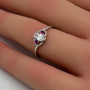 May include: A silver ring with a clear oval-shaped stone and two smaller pink stones on either side.