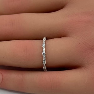 May include: A delicate silver ring with alternating round and baguette-cut clear stones. The ring is displayed on a hand with manicured nails, set against a soft, blurred background. The ring's design is simple yet elegant.