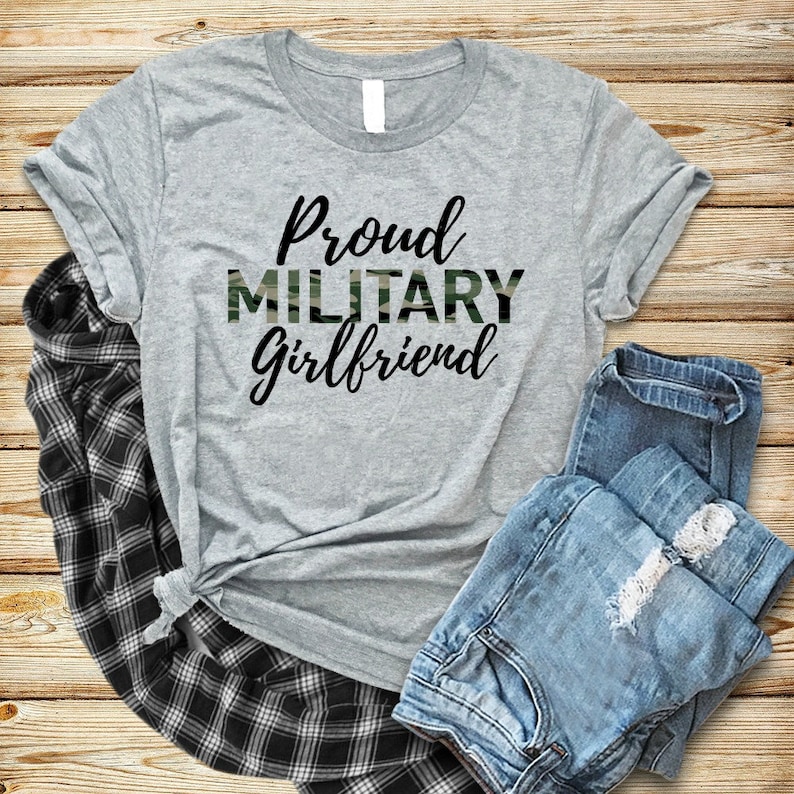 May include: Gray t-shirt with the text "Proud Military Girlfriend" in black lettering. The word "Military" is printed in a camouflage pattern.