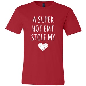 May include: Red t-shirt with white text that reads "A super hot EMT stole my" with a white heart drawn below the text.