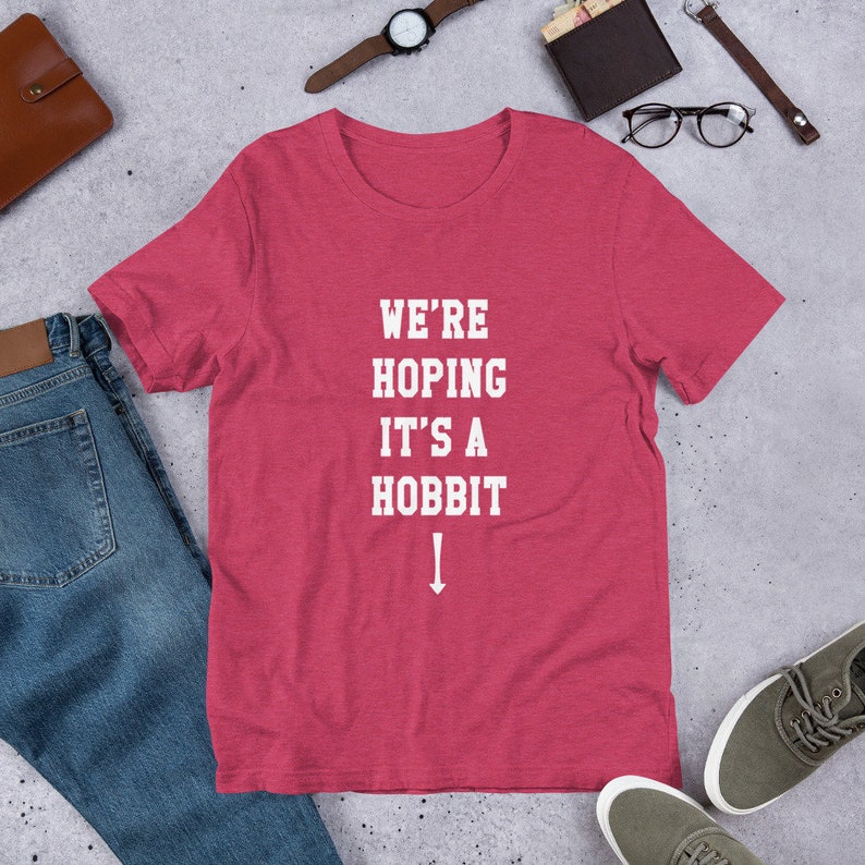 Może przedstawiać: Czerwony t-shirt w melanż z białym napisem "We're Hoping It's A Hobbit" i strzałką skierowaną w d&oacute;ł.