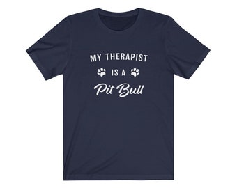 Op de afbeelding: Donkerblauw T-shirt met witte tekst die "My therapist is a Pit Bull" luidt, met twee poot afdrukken aan weerszijden van het woord "is".