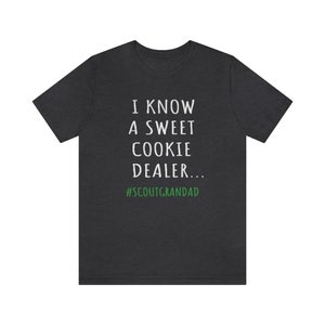 Op de afbeelding: Donkergrijs T-shirt met witte tekst die "Ik ken een lieve koekjesverkoper... #ScoutGrandad" in groen zegt.