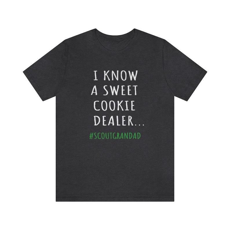 Op de afbeelding: Donkergrijs T-shirt met witte tekst die "Ik ken een lieve koekjesverkoper... #ScoutGrandad" in groen zegt.