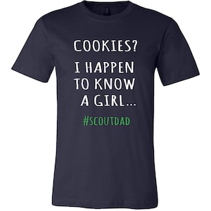May include: Navy blue t-shirt featuring the text "COOKIES? I HAPPEN TO KNOW A GIRL..." in white, with the hashtag "#SCOUTDAD" in green. A casual style t-shirt.